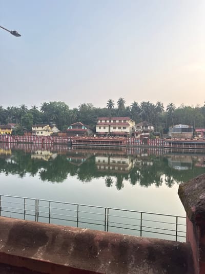 This image displays the Koti Teertha sacred pond in Gokarna, India, viewed across the water toward the eastern bank under a light, hazy sky.

In the immediate foreground, a weathered reddish-brown concrete barrier or ledge occupies the bottom segment, supporting a silver-colored metal railing composed of three horizontal bars that spans horizontally across the frame. A portion of a matching reddish-brown concrete post is visible at the far right.

The midground features a calm body of greenish-grey water, clearly reflecting the buildings, vegetation, and sky from the opposing bank.

On the far bank, a series of structures line the water's edge. Immediately bordering the water is a low-rise platform or retaining wall characterized by numerous vertical pillars painted with alternating red and white stripes. Behind this, several smaller buildings with dark, possibly tiled, roofs are visible. Further inland, a cluster of multi-story buildings includes a prominent cream-colored, three-story structure with a red tiled roof and numerous windows. Adjacent and further along the bank are other buildings of varying colors, including yellow, light-colored, and blue, some with indiscernible signage. Dense tropical vegetation, primarily palm trees, forms a lush green backdrop behind and between these structures.

The sky, occupying the upper third of the image, is a light, hazy blue-white. A dark grey street light fixture mounted on a pole extends into the upper-left portion of the sky. No individuals, specific actions, or interactions are discernible.