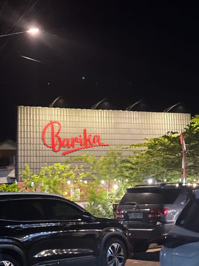 The image shows the front of a building in Lambaro, Indonesia, at night. The building has a sign that reads "Barika" in red cursive font. The building is covered with greenery and string lights. Two vehicles are parked in front of the building. A street light is visible in the background.
