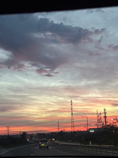 A vibrant sunset sky dominates the frame, transitioning from dark, heavy clouds on the left to a bright gradient of orange, pink, and yellow bands on the right. The scene is captured from a moving vehicle, with a road and blurred roadside vegetation visible along the bottom. Tall power line towers and other infrastructure punctuate the skyline, silhouetted against the colorful sky. A single car is visible on the road ahead, adding a sense of scale and movement.
