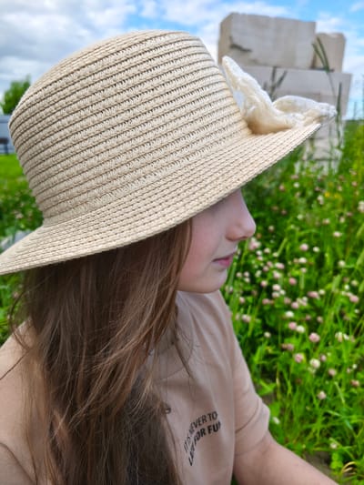 A young girl with long brown hair wears a straw hat decorated with a white fabric bow. She is looking to the right, her face partially obscured by the hat's brim. She is wearing a tan t-shirt with text that reads "IT IS NEVER TO LATE FOR FUN". She is outdoors in a grassy area with small pink and white flowers. In the background, there are large, light-colored, abstract stone sculptures. The sky is visible with some clouds.