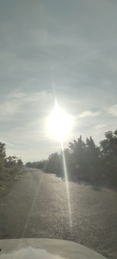The image depicts a road through a rural or semi-rural area, likely in Sumbokrayek, Indonesia, under a bright, overcast sky with the sun shining intensely through. The road is paved and appears to be a two-lane street. Dense vegetation, including trees and bushes, lines both sides of the road, with some scattered debris visible on the left shoulder. The sun's glare creates a strong lens flare, a vertical beam of light extending downwards from the sun, which is positioned high in the center of the frame. There are a few indistinct figures on motorcycles in the distance on the road. The foreground shows the hood of a vehicle, suggesting the image was taken from a car. The overall impression is a journey through a sun-drenched, green landscape.