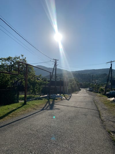 This media file depicts a sun-drenched, rural road in Aguy-Shapsug, Russia. The perspective is from a low angle, looking down a paved road that winds through a valley. The sun is prominently featured in the upper left quadrant, casting a bright glare and lens flare across the image. Power lines run along the side of the road, supported by wooden poles, indicating a settled area.

The scene is set against a backdrop of lush green hills and mountains, partially shrouded in a light haze, suggesting a warm, possibly humid, day. Trees and vegetation line the sides of the road, with some fences and what appear to be simple structures or homes visible. The road itself is asphalt, appearing somewhat worn, with gravel on the shoulders. Shadows are cast by the power poles and a lone tree, stretching across the road and grass.

There are no people visible in the image. The activity suggested is that of a quiet, possibly early morning or late afternoon, rural setting. The bright sunlight and clear blue sky point towards fair weather. The overall emotion evoked is one of serenity and natural beauty, typical of a countryside landscape bathed in sunlight. No text is visible in the image.
