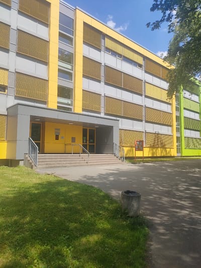 A modern building with a colorful facade stands under a bright blue sky with scattered clouds. The building features alternating bands of gray and vibrant yellow on its upper floors, punctuated by horizontal window blinds that are also yellow. The lower section of the building is a darker gray, leading to a bright yellow entrance area. This entrance is accessed by a wide set of concrete stairs with metal handrails. Two doors, one with glass and another appearing to be a solid yellow door with a mail slot, are visible in the entrance. There are signs on the yellow doors, one appearing to be a white rectangular sign with some text and a red graphic, and another smaller, red sign. A red display board stands to the right of the entrance. To the right of the main building section, the facade transitions to bright green, also with gray sections and yellow blinds. The area in front of the building is a paved walkway and a grassy lawn. A concrete trash receptacle with a black liner sits on the edge of the lawn near the walkway. Sunlight casts dappled shadows on the pavement and grass, suggesting it is a sunny day, likely late morning or early afternoon. The overall scene is clean and orderly, typical of a public or institutional building, possibly a school or office. The location context is Chemnitz, Germany.