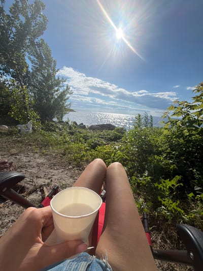 A person is sitting outdoors, holding a white paper cup of light-colored liquid. They are wearing denim shorts. The person is seated in a red chair with black armrests, outdoors. The background shows a body of water, likely a lake or river, with sunlight glinting off its surface. There are trees and green vegetation surrounding the area, with some rocks near the water's edge. The sky is blue with scattered clouds, and the sun is shining brightly. The scene appears to be a peaceful outdoor setting, possibly a picnic or leisure spot. The image does not contain any elements that would indicate it is located in Kozarovychi, Ukraine.