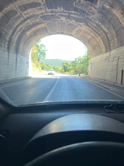 The image depicts the interior of a vehicle looking out through the windshield towards the exit of a concrete tunnel. The tunnel's arched ceiling, constructed from textured concrete with visible rebar patterns and staining, dominates the upper portion of the view. The tunnel walls are made of rough, light-colored concrete blocks. A paved road, marked with double white lines, leads out of the tunnel into bright daylight. Sunlight illuminates a white car positioned on the road ahead and beyond, a lush green landscape with trees and rolling hills under a clear sky. The foreground shows a portion of the car's dashboard and steering wheel, out of focus. The location is identified as Kruče - Kërruçi, Montenegro.