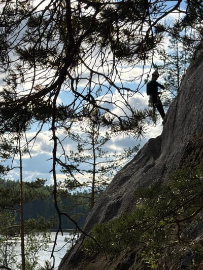 The image depicts a silhouette of a rock climber ascending a steep, grey rock face. The climber, dressed in dark clothing, is positioned on the upper right side of the rock. They are secured by a climbing rope, which is visible as it hangs down and coils slightly around their waist. The climber is facing to the left, appearing to be in the process of moving upwards.
In the foreground, silhouetted pine branches with sharp needles create a dark, framing effect across the upper portion of the image. The branches are dense and create a dappled pattern of light and shadow.
The background reveals a serene natural landscape. Below the rock face and to the left, a body of water, likely a lake or river, is visible. The water reflects the light and appears calm. Beyond the water, a dense forest of evergreen trees stretches across the horizon, indicating a wilderness setting. The sky above is a mix of blue with scattered, soft white clouds, suggesting it is daytime.
The scene is consistent with a natural outdoor climbing location. While the specific city of Ruch'i, Russia is mentioned in the prompt, the visual information in the image alone does not provide any direct evidence to confirm this location. However, the presence of a lake, extensive forest, and rocky terrain is characteristic of many natural areas found in Russia.