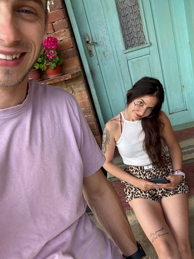 The image shows a man and a woman sitting on a porch in front of a turquoise door. The man is in the foreground, his face taking up the left side of the frame. He is smiling and wearing a light purple t-shirt. Behind him, on a shelf attached to a brick wall, is a potted pink flower. To the right of the man, the woman is seated, looking towards the camera with a slight tilt of her head. She has long dark hair, is wearing glasses, a white tank top, and leopard-print shorts. She is holding a smartphone. A tattoo is visible on her left arm, and another tattoo with text is visible on her right thigh. The floor of the porch is covered by a rug with a striped pattern. The overall setting appears to be residential, possibly in the village of Borintsi, Bulgaria, given the architectural style of the brickwork and the door.