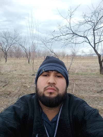A man wearing a dark beanie and black jacket poses for a selfie. He has a full beard and a patterned shirt visible under the jacket. The background is an overcast landscape with many bare trees scattered across a field of dry grass and dirt. The scene suggests a cold or early spring day.