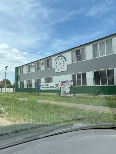 This media file shows a building with several windows, likely a school or training center. The building is clad in grey and green siding. In front of the building, there's a green metal fence and a grassy area. Several posters are visible attached to the fence. One poster reads "ВЫПУСКАЕМ ПРОФЕССИОНАЛОВ!" (We are graduating professionals!) and another features colorful shapes with the text "ПРОФЕССИОНАЛИТЕ 2022" (Professionalism 2022). Further to the right, a poster in Russian advertises "КРАСНОДАРСКИЙ НАБОР В ВОЙСКА БЕСПИЛОТНЫХ СИСТЕМ" (Krasnodarsky recruitment into drone systems troops), with figures of money (63.4 million rubles) and a phone number. Another poster indicates "85 лет СТУ" (85 years of study/student). The scene is outdoors, under a bright blue sky with scattered clouds. A paved path runs alongside the grassy area. The photo is taken from the perspective of someone inside a vehicle, as indicated by the partial view of a car's windshield and wiper on the right side of the frame. The time of day appears to be daytime, given the bright sky. The weather seems to be pleasant.