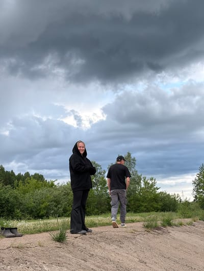 The image shows two people on a dirt road with green vegetation on either side and a cloudy sky above.
The person closer to the camera is wearing a black hooded jacket and black pants. They are smiling and looking at the camera.
The person further away is wearing a black shirt, grey pants, a black baseball cap, and sunglasses. They are facing away from the camera.
In the background, there is a power line visible among the trees.
Based on information available, it is not possible to definitively identify the specific location as Nizhnii Vishur, Russia, without further evidence.