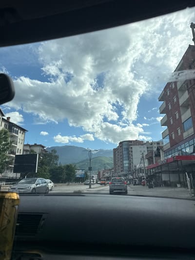 The image shows a view from inside a car looking out onto a street in the city of Pejë, Kosovo. The sky is visible with fluffy white clouds against a blue backdrop. Buildings line both sides of the street, ranging from low-rise residential structures to taller commercial buildings. In the distance, mountains are visible. Cars are present on the street, including a grey Volvo SUV in front of the vehicle from which the photo was taken. There are also other cars parked along the sides of the street. Various street signs and utility poles are present.