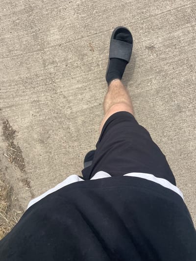 The image shows a person's legs from the thigh down, walking on a concrete surface. The person is wearing black shorts with a white stripe near the hem, black socks, and black slide sandals. The concrete appears to be outdoor pavement, possibly a sidewalk or path, with some dry grass and debris on the left side of the frame. The lighting suggests it is daytime. The image does not provide any specific visual cues that would allow for precise location identification within Diepenbeek, Belgium.