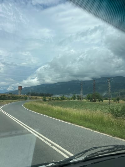 The image presents a view from inside a vehicle, looking out at a rural or mountainous landscape under an overcast sky. A paved road curves into the distance on the right side of the frame, marked by white double solid lines on its right edge and a single white line bordering a grassy shoulder on the left.
To the left of the road, a wide expanse of green, wild grass stretches out, interspersed with small yellow wildflowers. Further into the middle ground, several tall metal electricity pylons stand prominently, carrying numerous power lines that traverse the scene horizontally. Beyond these structures, dark, imposing mountains rise towards the horizon, their peaks partially veiled by clouds or atmospheric haze. The sky is predominantly cloudy, with shades of grey and white, suggesting an overcast day.
A distinctive reddish-brown building, possibly a water tower or an old industrial structure, is visible near the base of the mountains, amidst some scattered trees and vegetation. The top right corner of the image shows a partial view of the vehicle's interior, including windshield wipers, while the top left corner displays a textured pattern, likely from the vehicle's ceiling or sunshade.
The scene depicts a cloudy daytime journey through a vast, natural environment, likely in the countryside. There is no visible text in the image. The location is Vetren, Bulgaria.