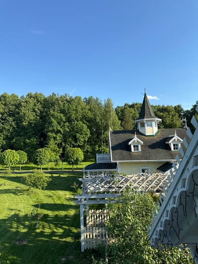 This media file captures a serene, well-maintained landscape featuring a distinctive house and garden under a clear blue sky, likely in Nadezhda, Russia. The scene is dominated by a two-story house on the right, painted light yellow with a dark shingled roof. Its architecture is notable for its intricate white trim, characteristic of Victorian or Gingerbread styles, featuring decorative dormer windows and a prominent conical turret with a finial at its apex. A smaller, lighter section with windows is visible on the turret, possibly a belvedere.

In the foreground, a vibrant green lawn extends, dappled with shadows from unseen structures or trees. Several neatly pruned, spherical bushes are aligned along the lawn's edge. A large white wooden lattice structure, resembling a pergola or arbor, frames the right side of the image, partially obscuring the house and extending towards dense green foliage. String lights are visible underneath this structure. A segment of a decorative white wooden fence or gate is also seen to the far right.

Behind the house and garden, a dense line of tall, mature green trees forms a lush natural backdrop, suggesting a forested area or a large park. The sky above is a brilliant, cloudless blue, indicating clear weather. The strong sunlight and defined shadows on the grass suggest it is late morning or early afternoon. No people or specific activities are visible, presenting a peaceful, static view of an architectural and natural setting. There is no visible text in the image.