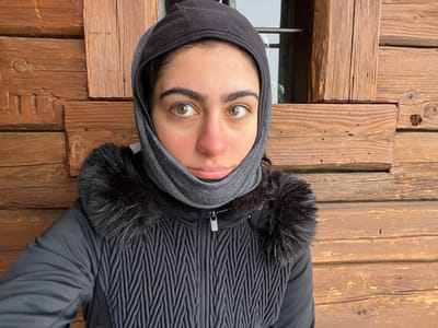 A young adult female is depicted outdoors, positioned against a rustic wooden building facade in Valtournenche, Italy. She wears a black hooded jacket with a quilted chest panel and a black faux fur trim around the collar, zipped up at the front. A gray balaclava covers her head and neck, leaving her face exposed. Her light-colored eyes are directed towards the viewer, and her cheeks and nose show a faint reddish hue, indicative of cold conditions. The background consists of horizontally aligned, weathered wooden planks or logs. In the upper right, a window opening with a single horizontal metal bar is partially visible.