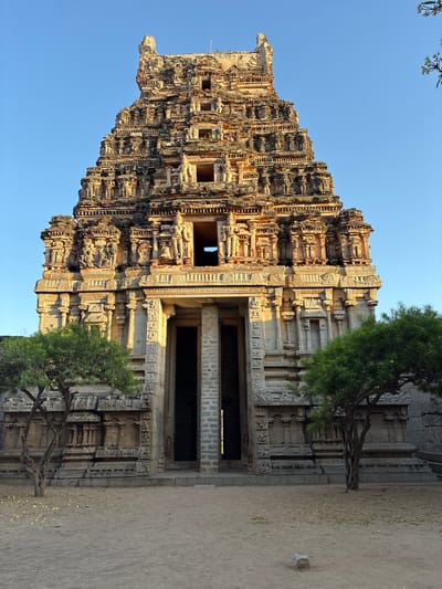 The image captures the Hazara Rama Temple in Kamalapura, India. The temple's towering gopuram, adorned with intricate carvings, dominates the frame. The temple entrance is flanked by carved pillars and stone walls. Two trees with green foliage frame the lower part of the structure. The ground is visible, a tan color. The sky above the temple is a clear blue.