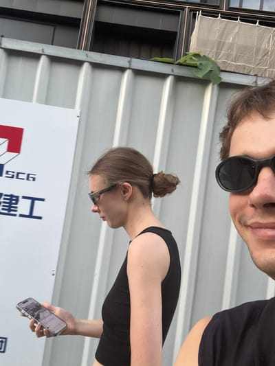 The image, presented sideways and requiring a 90-degree clockwise rotation for proper viewing, captures two individuals in a bright, likely outdoor setting.

In the foreground, a man, partially visible from the forehead to the chin, appears to be taking a selfie. He wears black sunglasses that obscure his eyes and has short brown hair with light facial stubble. He is dressed in a dark t-shirt.

Beside him, a woman is seen in profile, facing towards the right side of the frame. She has brown hair neatly tied in a bun and wears clear-lensed eyeglasses with dark frames. She is dressed in a black sleeveless top. Her right hand holds a smartphone, possibly an iPhone, which she is actively looking at; the screen displays what appears to be a messaging app with text and an image.

The background predominantly features a light grey, corrugated metal wall or cladding, suggesting an industrial or construction environment. To the far left, a dark building structure with some reflective material is visible, along with a small cluster of green leaves or a vine.

A large white sign dominates the lower portion of the image. It features a red and white geometric logo, the English letters "SCG," and partially visible Chinese characters that read "工程" (gōngchéng), which translates to "engineering" or "project." The bright lighting indicates it is daytime. Neither individual displays overt emotions, with the woman focused on her device. Given the "SCG" and "工程" text and the Putuo District context, the location likely pertains to an engineering or construction project.