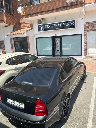 This image captures a street scene in Villa del Prado, Spain, on a sunny day. A black sedan is parked in the foreground, its rear end facing the camera. The license plate reads "2034 DSF". To the left of the sedan, a portion of a white SUV is visible. Behind the vehicles, a storefront with a large window displays "DESIRÈE VALVERDE Hair Beauty" on a prominent sign. An LG air conditioning unit is mounted above the storefront. The building is constructed with brick and white plaster. Sunlight casts sharp shadows on the asphalt parking lot and the tiled pavement in front of the building.