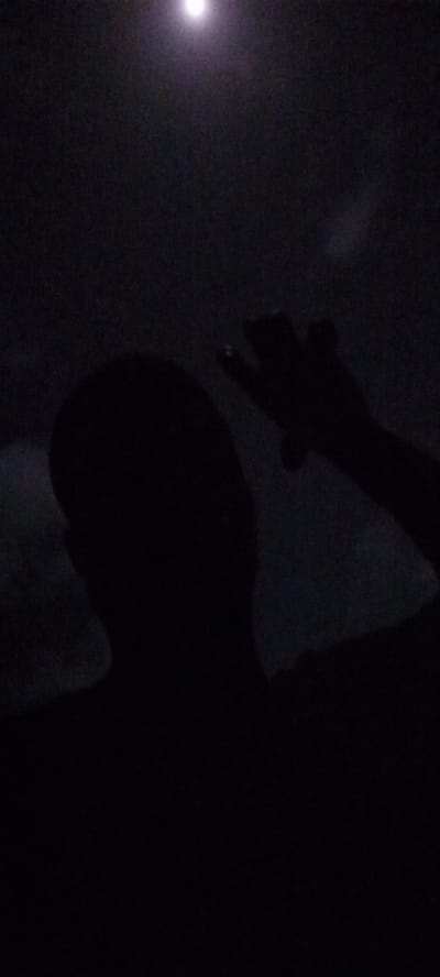 The image is a low-light, silhouetted portrait. It appears to be taken at night, possibly under moonlight or weak artificial lighting. The primary subject is a person, visible only as a dark silhouette. Their head and upper torso are positioned in the lower-left to center of the frame. An arm is raised, with the hand slightly obscured but visible, possibly gesturing or reaching. The background is a dark, mottled expanse, hinting at clouds or other indistinct shapes. The location is Ilogbo Eremi, Nigeria. No specific details or objects are discernible that could provide forensic information about actions, interactions, or precise environmental conditions.