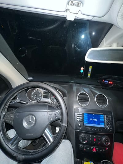 The image depicts the interior of a Mercedes-Benz vehicle, viewed from the driver's perspective, at night. The location is specified as Piperkov Chiflik, Bulgaria.

A person is seated in the driver's seat, their legs clad in light grey sweatpants visible below the steering wheel. The multi-function steering wheel, featuring a Mercedes-Benz logo, is wrapped in a black aftermarket cover. The instrument cluster behind the steering wheel indicates a vehicle speed of 0 km/h and an engine RPM of 0, suggesting the vehicle is stationary.

The center console features two circular air vents above a multimedia display showing "99.9 MHz" and "Radio FM2," indicating the audio system is active. Below the display are climate control dials and various buttons, including a red triangular hazard light button. The footwell on the driver's side shows a perforated metallic accelerator pedal. A red object, possibly a garment or bag, is visible on the passenger side floor or seat area. The windshield appears dusty or smudged, with reflections visible on its surface.

Outside the vehicle, viewed through the windshield against a dark night sky, are two distinct road signs on the left: a blue circular sign with a white arrow (likely 'ahead only') and a red circular sign with a white horizontal bar (likely 'no entry' or 'stop'). A bright white light source, possibly a streetlight or oncoming headlight, is visible further down the road. On the right side of the exterior view, partial details of another dark-colored vehicle with illuminated red lights are discernible. An overhead light panel with controls is visible above the windshield.
