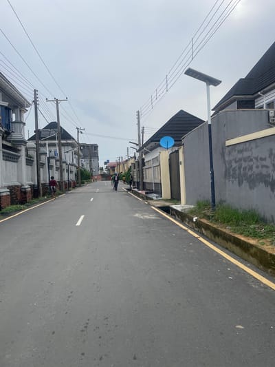 This is a street-level, eye-level, full-shot photograph of a paved road in Rumuodara, Nigeria, under an overcast sky. The road is bordered by a raised concrete curb with overgrown grass and a painted yellow line along the edge. On the left, a white-walled building with a red-brick base and decorative pillars lines the street. On the right, a gray-walled building with a dark tiled roof is visible. Several utility poles with electrical wires stretch across the frame.
There are several people on the road. In the foreground on the left, a person wearing a red shirt and dark shorts walks away from the camera. Further down the road, a group of individuals are visible. One person is wearing a green shirt and black shorts, and another is wearing a black shirt with the number 11 in white. Two individuals appear to be pushing a cart. The architecture of the buildings suggests a residential area with modern design elements. The overall scene depicts a typical street in a Nigerian urban or suburban environment.