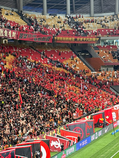 The image depicts a crowded stadium in Longxing, China. The stadium seating is divided into sections, with the left side predominantly yellow and the right side primarily red. A dense crowd of people occupies the seats, with many wearing red and black attire, suggesting team colors. Banners with Chinese characters and logos are displayed throughout the stadium. A green field is visible at the top right corner of the image. The stadium's architecture includes metal supports and walls.