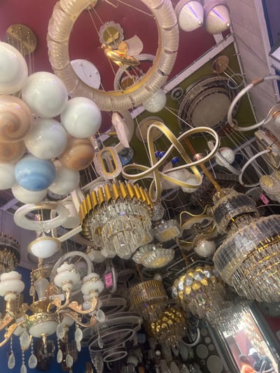 The image displays a cluttered, upward-angled view inside a retail lighting store in Igbede, Nigeria. The scene is dominated by a dense array of chandeliers and various light fixtures hanging from a ceiling painted in sections of red, olive green, and white.
Numerous chandeliers are visible, including multi-tiered crystal designs with gold-toned metal accents, some partially wrapped in protective clear plastic. Modern light fixtures are interspersed, featuring spherical glass shades in white, marbled brown, and light blue. Other contemporary designs include ring-shaped LED fixtures, abstract intertwined gold and white ribbon-like structures, and smaller flush-mount or semi-flush-mount units with frosted or textured glass. Wires and suspension hardware are visible throughout the ceiling.
In the lower right portion of the image, a reflection is observed in a dark, vertical surface, possibly a mirror or reflective pane. This reflection shows the partial figure of an individual wearing a white cap and an orange shirt, suggesting the presence of a person (customer or staff) within the retail space. No direct interaction or specific action is discernible. The overall environment appears to be a commercial showroom for residential and decorative lighting.