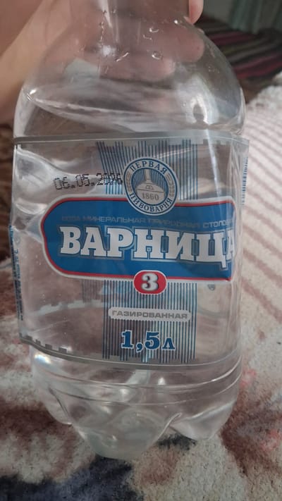 The image is a close-up shot of a plastic bottle of mineral water. The label is predominantly blue and white, with Cyrillic text. The brand name "ВАРНИЦА" (Varnitsa) is prominently displayed in large white letters. Above the brand name, there is a circular emblem with the text "ПЕРВАЯ ПИВОВАРНЯ" (First Brewery) and the year "1860". Below the brand name, it states "ГАЗИРОВАННАЯ" (Carbonated) and "1,5л" (1.5L). A red circle with the number "3" is located within the label. The bottle is filled with clear liquid. The background appears to be a textured surface, possibly fabric, in muted colors. There is no indication of the city of Beloci, Moldova, in the image.