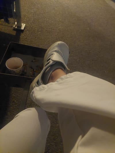 A high-angle, close-up shot shows a person's leg and foot in Altdorf, Germany, positioned next to an ashtray. The person wears white pants and a light gray athletic shoe with velcro straps. The ashtray is a dark gray metal container filled with cigarette butts. Beside the cigarette butts is a partially empty paper cup. The ground around the ashtray appears to be paved. A small section of a blue metal structure is visible in the lower left corner. The scene is dimly lit, possibly at night.