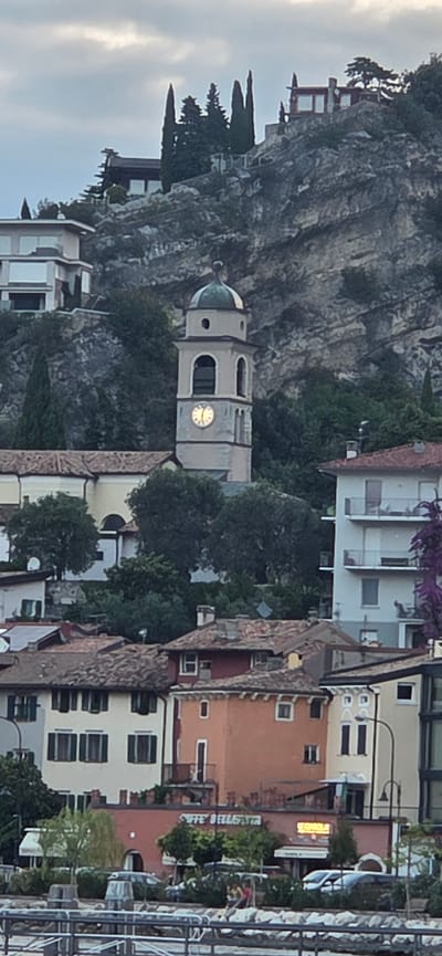 The image shows a town nestled against a rocky hillside in Torbole, Italy. A prominent clock tower with a green dome stands tall, its clock face illuminated. Below the tower, a cluster of buildings with red-tiled roofs are visible. The buildings range in color from light yellow and beige to orange and red. Trees are scattered throughout the scene, with some lining the slopes of the hill and others in front of the buildings. In the foreground, a body of water is visible, with a rocky embankment and a railing suggesting a promenade or walkway. The sky is overcast with diffused sunlight.