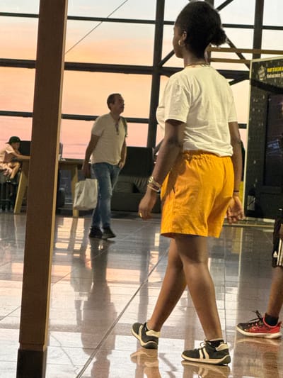 The image depicts an indoor public space, likely an airport terminal, with large windows reflecting a sunset sky in warm orange and pink hues. A woman with dark hair pulled back is walking away from the camera, wearing a white t-shirt and bright orange shorts. She has on black and white sneakers and several bracelets on her left wrist. To her left, a man in a light-colored polo shirt and blue jeans is walking in the opposite direction, carrying a white tote bag. He has a backpack slung over one shoulder. In the background, a young person wearing a light-colored shirt and a dark baseball cap is seated at a counter. The floor is reflective, showing the light from the windows. The overall scene suggests a travel hub during late afternoon or early evening. The location is identified as Zaventem, Belgium, which is the location of Brussels Airport.