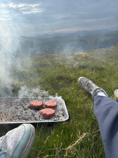 A high-angle shot depicts a barbecue grill with four uncooked hamburger patties emitting smoke. The grill sits on grass, with two pairs of legs and sneakers visible on either side of the grill. In the background, a landscape scene displays hazy mountains and sky.