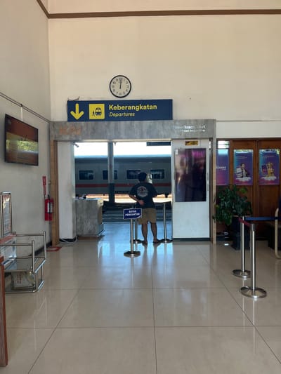 The image depicts the interior of a train station in Desa Tambakreja, Indonesia. A clock on the wall reads approximately 12:05. A sign with a yellow arrow pointing down and a depiction of a train indicates "Keberangkatan Departures." To the right, a sign reads "Pemeriksaan Tiket Ticket Inspection." A man in a dark t-shirt and shorts is standing facing a train, with his hands behind his back. A fire extinguisher is mounted on the wall to the left. The floor is tiled and reflective. In the background, a train is visible through an open doorway. Posters are displayed on the right wall, one of which mentions "Access by KAI."