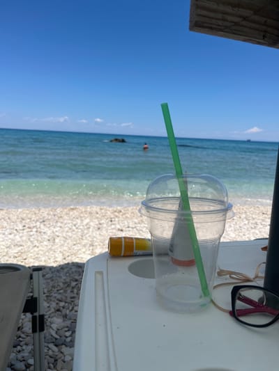 An eye-level medium shot shows a clear plastic cup with a green straw on a white table at a beach in Elaties Municipal Unit, Greece. The cup is partially filled with a clear liquid and condensation. To the left of the cup, a yellow tube of sunscreen is visible. To the right of the cup, a pair of black-framed glasses with pink arms rests on a thin brown string. In the background, the turquoise ocean meets a pebble beach under a clear blue sky. A person can be seen swimming in the ocean in the distance. A large wooden beam from a beach umbrella is visible in the upper right corner.