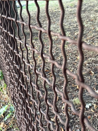 A close-up, slightly angled shot shows a rusty chain-link fence in Piatykhatky, Ukraine. The fence is made of intertwined wires with a wavy pattern, and the metal appears corroded. Behind the fence, the ground is covered in dry grass, fallen leaves, and a few sparse green plants. A tree trunk, also slightly out of focus, is visible to the left behind the fence. The overall impression is one of outdoor neglect or disuse.