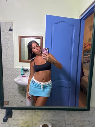 The image is a reflection in a bathroom mirror. The subject is a young woman with dark hair, wearing a black sports bra, and a blue, white, and yellow mini skirt. She is holding a smartphone with a pink leopard print case in her right hand, and taking a selfie. Her left hand is on her hip. She is smiling and puckering her lips for the photo. There is a white sink and counter to the left of her reflection. Above the sink is a small wooden shelf holding a bottle of lotion. There is a bathroom door painted in a vibrant blue color to her right. The door appears to be closed. The reflection in the mirror shows a room behind the woman with a bed and other furniture. The image appears to be taken in a bathroom in a residence in Montañita, Ecuador, with coordinates of latitude -1.825 and longitude -80.752.