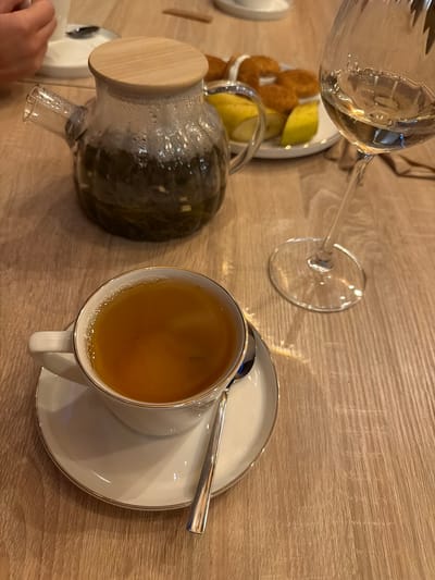 In Sharovechka, Ukraine, a cup of tea sits on a saucer on a wooden table, accompanied by a teapot and a glass of white wine. A plate of food, possibly pastries or appetizers, is visible in the background, along with a partial view of a person's hand. The scene suggests a relaxed moment of enjoyment, perhaps a break or a social gathering, in a comfortable setting.