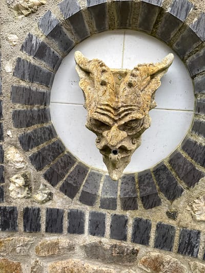 A weathered, light brown, gargoyle-like stone carving functions as a waterspout, mounted centrally on an outdoor wall in Stiperstones, United Kingdom. The carving depicts a stylized face with prominent brow ridges, upward-curving horn-like protrusions, and a dark circular metal spout protruding from its mouth area. A single visible screw secures the carving above its right horn. It is affixed to a circular panel of white square tiles, approximately five to six tiles in diameter, with visible grout lines. This tiled panel is framed by a radially arranged arch of dark grey, rectangular tiles, which exhibit vertical water streaks. Surrounding this arched frame, the wall consists of irregularly shaped natural stones, including lighter, possibly quartz or flint, fragments, all set in a light grey mortar. No individuals or dynamic actions are present in the scene.