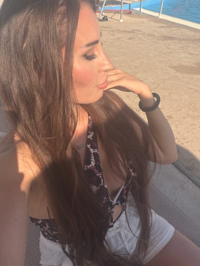 The image features a young woman lying on a lounge chair, presumably outdoors. She has long, dark brown hair that cascades over her shoulders and face. Her eyes are closed, and her head is tilted back, suggesting a relaxed or contemplative mood. She is wearing a bikini with a patterned top and white bottoms, and a thin silver necklace. Her right hand is raised, with fingers lightly touching her chin and lips.
The background shows a beige, textured surface, possibly a deck or patio, with dappled sunlight casting shadows. To the left, a glimpse of a swimming pool with blue water and metal railings is visible. The scene appears to be set on a sunny day, given the bright lighting and strong shadows. The overall impression is one of leisure and relaxation in a warm-weather location.
There is no visible text in the image.