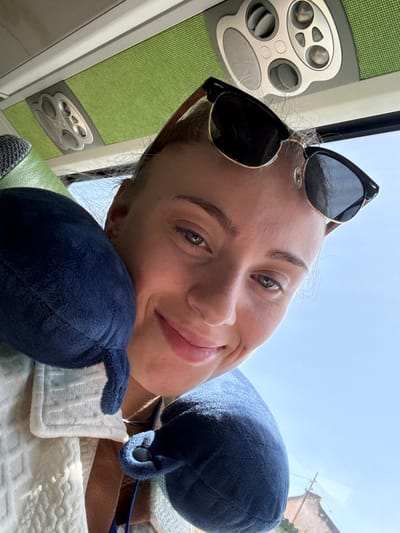 A woman is taking a selfie in what appears to be a mode of transportation, possibly a bus or train. She is wearing sunglasses and a blue travel pillow is supporting her head. Another blue travel pillow is visible in the foreground. The woman has a relaxed and happy expression.

The interior of the vehicle is visible, with green upholstery and a control panel featuring buttons and vents. The lighting suggests it is daytime, and the bright sky visible in the background hints at clear weather. The overall scene conveys a sense of comfort and travel. There is no visible text in the image. Given the location context of Faro, Portugal, it is possible she is traveling within or around this city.