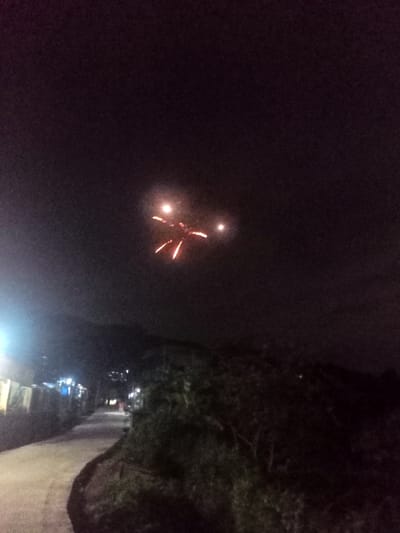 This night photograph, taken in Kecamatan Pangalengan, Indonesia, features a dark sky illuminated by a bright, luminous object. The object, possibly a type of firework or a drone with pyrotechnic elements, displays a central bright light, two additional bright points to its sides, and three distinct red-orange fiery trails descending from its core, giving it a unique, star-like or crab-like appearance. Below the sky, a dimly lit, unpaved road or path curves towards the left. On the left side of the path, several buildings are visible, illuminated by a few artificial streetlights. The right side of the path is bordered by dense trees and foliage. In the background, dark silhouettes of hills or mountains can be seen against the cloudy night sky. No people are visible in the image, and there is no discernible text. The overall scene conveys a quiet night setting with an unexpected visual display in the sky.