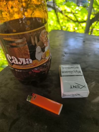 The image shows a close-up of a dark wooden table with a bottle of "SALA" beer, a pack of Kent cigarettes, and an orange BIC lighter. The beer bottle has a label with a drawing of a man with a beard, a barrel, and some fish hanging. Text on the label reads "БОЧКОВЕ ПИВО" and "SALA" in large letters, with "Вміст спирту 5,1% об." below. The cigarette pack is white and silver with the "KENT" logo and some text in Cyrillic script, possibly reading "КУРІННЯ ВПЛИВАЄ" (smoking affects). The BIC lighter is bright orange with the "BIC" logo visible.
The setting appears to be outdoors, with blurred green foliage and dappled sunlight suggesting it is daytime. The lighting and shadows imply it could be late morning or early afternoon. The overall impression is a casual, possibly relaxed, moment. The combination of beer, cigarettes, and a lighter suggests a break or leisure time. No people are directly depicted, but the context implies someone is present, likely the one taking the photo. The background foliage suggests a natural or garden-like environment, possibly a park or backyard.