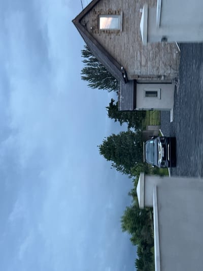 The image shows a side view of a modern stone-clad house with a dark-tiled roof and a light-colored modern extension. A black SUV is parked on a gravel driveway in front of the house. The sky is overcast and appears to be either dusk or a cloudy day. Trees and greenery are visible in the background, suggesting a suburban or rural setting. A metal gate is partially visible behind the parked car, leading to a grassy area. The overall scene is calm and residential. The location is identified as Unknown, Ireland.