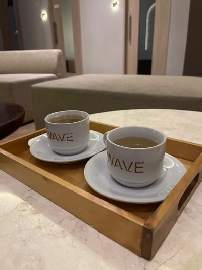 Two white ceramic cups with a gold-colored "WAVE" logo are placed on white saucers, which are arranged on a wooden tray. The tray sits on a marble-patterned table. In the background, a beige sofa and an ottoman are visible, along with doorways and interior lighting, suggesting an indoor setting that could be a lounge or reception area. The location is not discernable from the image provided.
