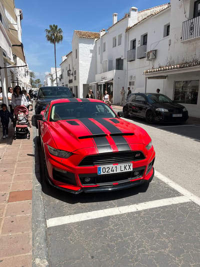 The image depicts a street scene in Nueva Andalucía, Spain, with a red Ford Mustang with black racing stripes parked on the right side of the road. The car's license plate reads "0241 LKX". A black car is parked behind the Mustang. The road is marked with white parking lines. On the left side of the road, there are white buildings with multiple levels, balconies, and air conditioning units. People are walking on the sidewalk, including a person pushing a stroller. A palm tree is visible in the background. Awnings extend from buildings on the left. The weather appears sunny.