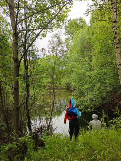 Two individuals are positioned on the grassy bank of a body of water, surrounded by dense green foliage and trees. The person in the foreground, facing away from the camera, is wearing a hooded jacket with a multi-colored, tie-dye pattern of red, blue, and white, along with dark pants and dark boots. They are holding a fishing rod, with the line extending into the water. To their right and slightly further back, a second person, also facing away, is wearing a light gray hooded sweatshirt and appears to be seated. The environment suggests a natural, possibly wild or semi-wild area, with the water appearing murky and reflecting the overcast sky and surrounding vegetation. The lighting indicates an outdoor scene, likely during daylight hours under cloudy conditions. The location is identified as Medvedyovka, Russia.