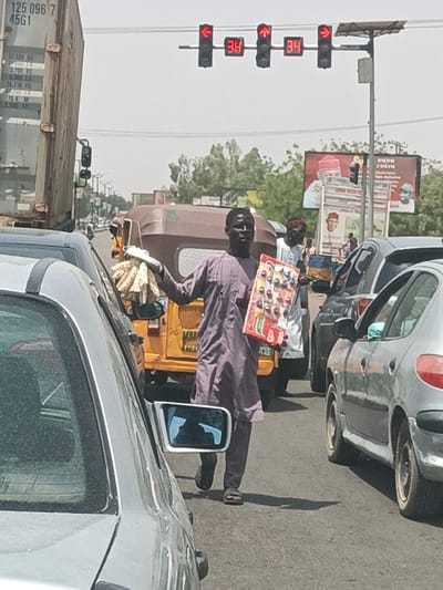 The image depicts a daylight street scene in Hotoro, Nigeria, focusing on a traffic intersection with stopped vehicles. A dark-skinned male street vendor, wearing a long-sleeved purple tunic and black sandals, stands facing forward in the foreground, positioned between two lanes of stopped cars. He holds a bundle of light-colored, rolled items in his outstretched left hand and a rectangular red display board with multiple colorful small items (watches or toys) in his right hand.
Above the street, a structure supports four active traffic lights, each displaying a red signal and the digital number "34." The outer two lights show red turn arrows (left and right), while the inner two show red upward arrows. A small solar panel is attached to the right side of the traffic light pole.
Vehicles populate the roadway. In the far left lane, the rear of a large beige cargo container truck (labeled "125 096 7 45G1") is visible. Directly behind the main vendor, a yellow-brown three-wheeled auto-rickshaw is stopped. A silver-grey car occupies the immediate left foreground, with its side mirror visible. In the right lane, a silver-grey hatchback (resembling a Peugeot 206) is stopped. Further back, another male vendor in a white tunic and a red-patterned cap is visible between cars, holding a blue and yellow object.
In the mid-ground, a large billboard featuring images of political figures and text, including "ABUBAKAR" and "BABA ZAHAR," stands near the road. The environment includes an asphalt road surface, clear skies with a slight haze, and green trees lining the distant road. Strong shadows are cast on the asphalt, indicating bright sunlight.