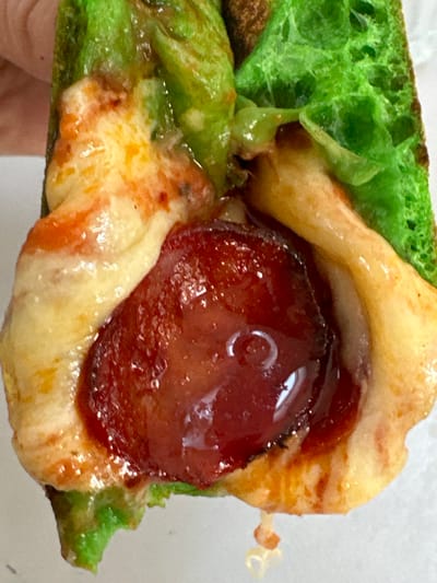 The image shows a close-up of a hot dog bun that has been dyed green, filled with melted cheese, sauce, and a slice of pepperoni. The bun appears to be slightly charred in places. The food is being held by a person's hand, though only a portion of the hand is visible. The background is a plain white surface. There is no discernible location in Moortown, Ireland, or any other specific environment.