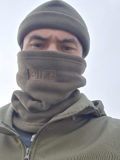 A close-up image captures an individual, from the upper chest to the top of the head, against a featureless light gray background indicative of an overcast sky. The person is wearing a drab green fleece beanie and a matching drab green fleece neck gaiter pulled up to obscure the nose, mouth, and chin. A reversed, embroidered mark, resembling '5.11', is visible on the front of the neck gaiter. The individual's dark eyes and eyebrows are visible, directed towards the viewer. They are also wearing a partially unzipped drab green fleece jacket, revealing a dark undershirt. The scene is situated outdoors in Egizbulak, Uzbekistan.