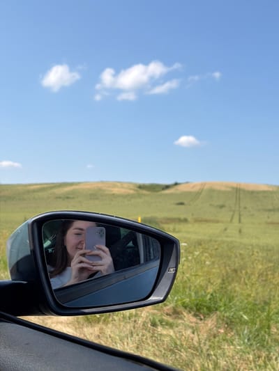 In the foreground, a car's side mirror reflects a young woman holding a purple phone and taking a selfie. Her eyes are partially closed, and she is smiling. The background of the mirror shows the interior of a car. Outside the mirror, a vast, grassy field stretches towards rolling hills under a clear blue sky with scattered white clouds. The field has visible tracks, suggesting agricultural activity. The scene is set in the countryside, possibly in the Astrashitskagaradokski rural council, Belarus, given the landscape.
